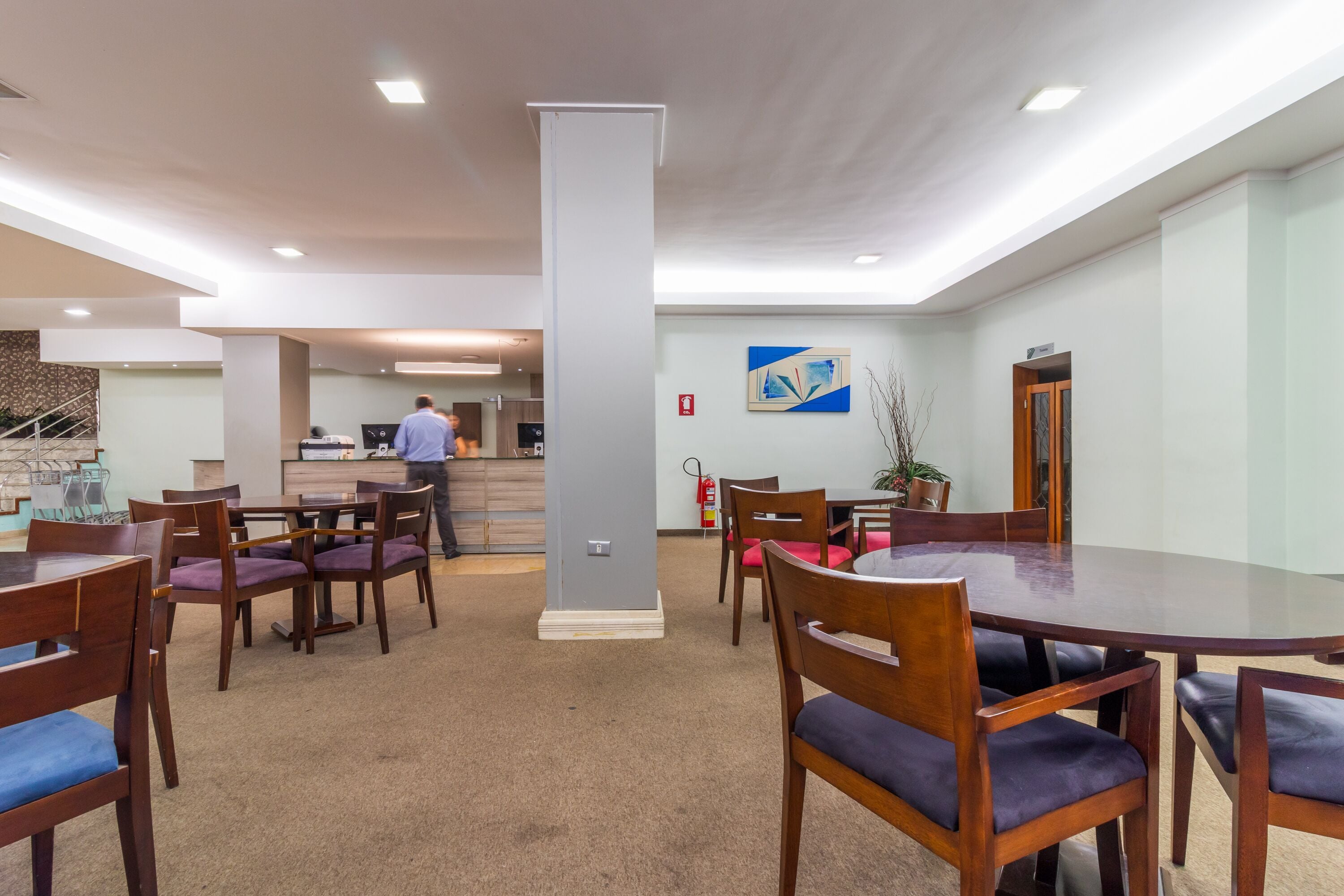 Un comedor moderno con mesas y sillas de madera, un pilar central y una cocina al fondo.