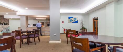 Un comedor moderno con mesas y sillas de madera, un pilar central y una cocina al fondo.