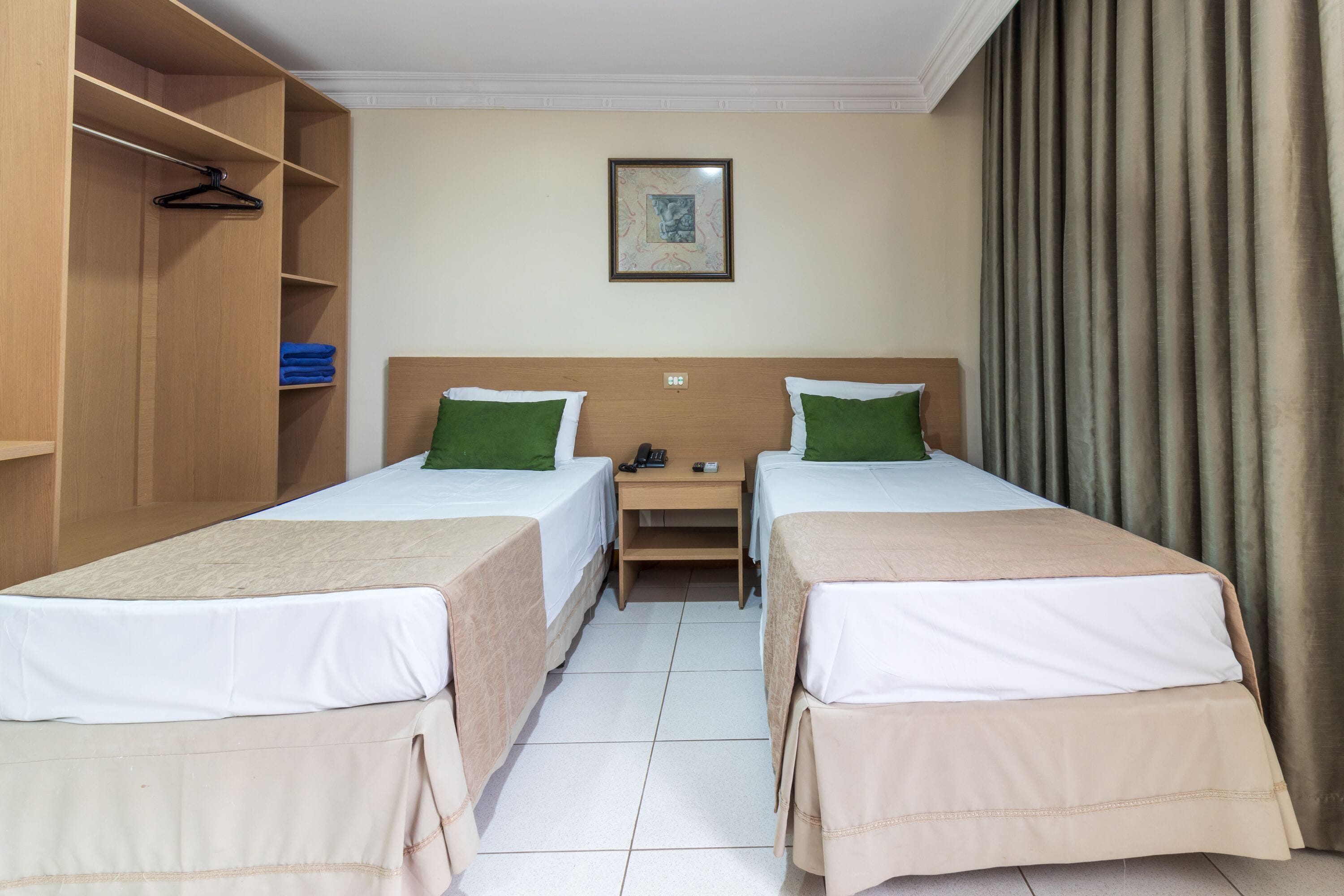 Una habitación de hotel con dos camas individuales, un armario de madera, una mesita con un teléfono y un control remoto, y un cuadro en la pared.