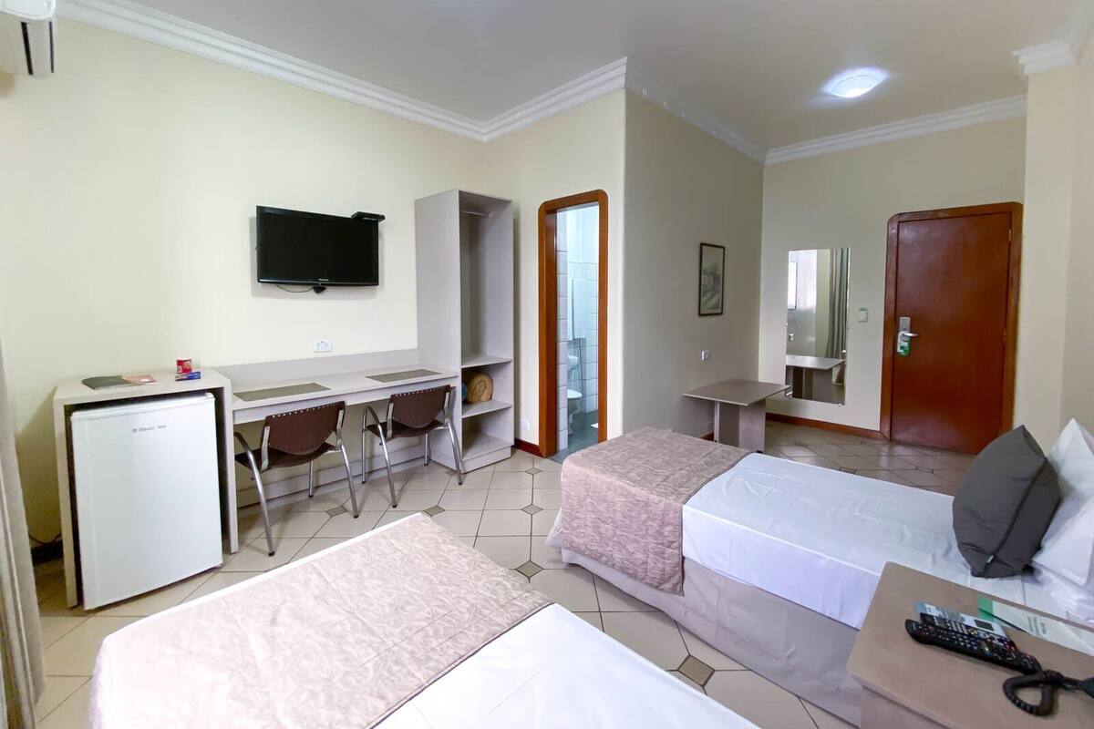 Ett hotellrum med två sängar, ett skrivbord, en tv, ett kylskåp och en spegel på badrummet.