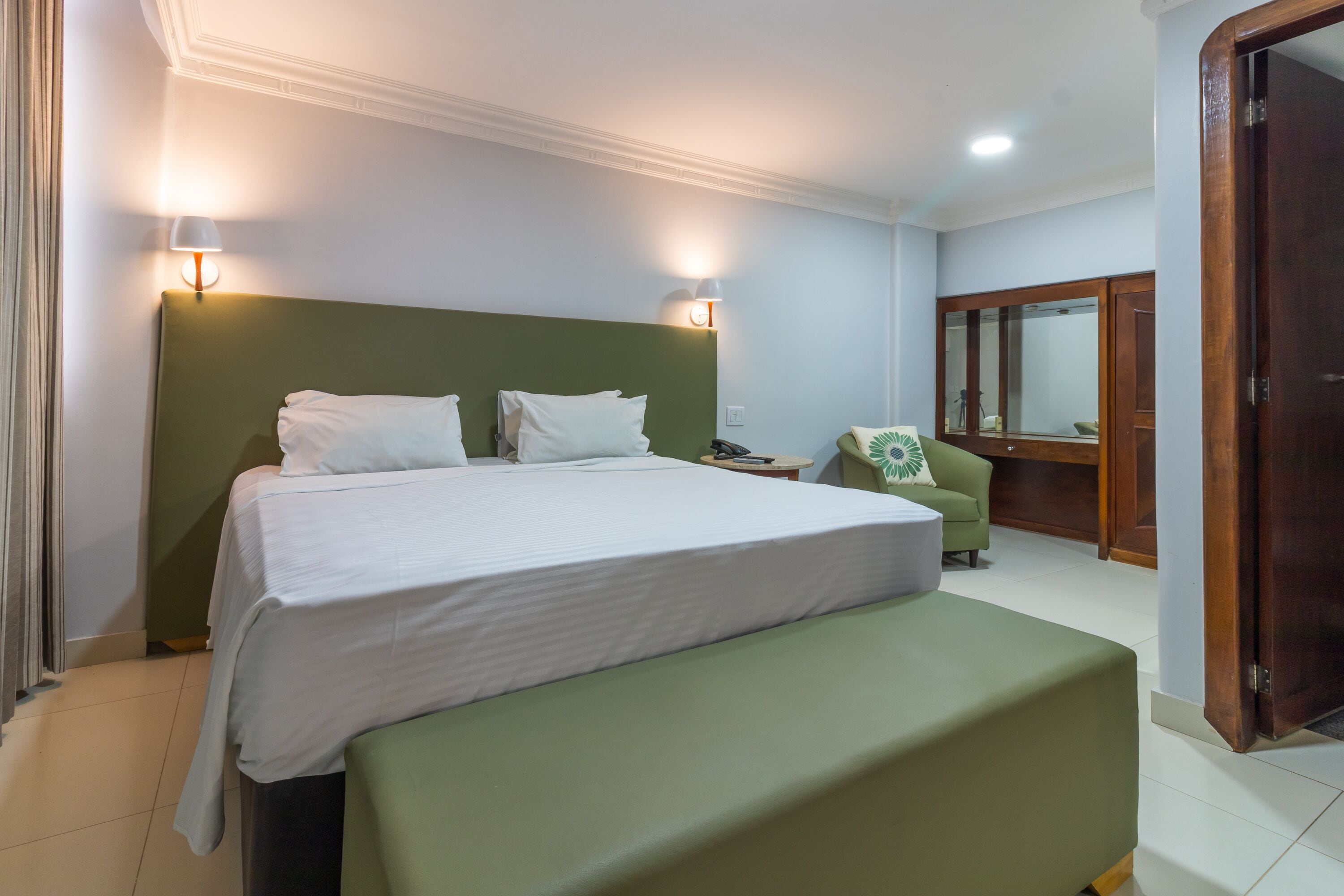 A hotel room with a large bed, a green bench, a small table, a chair, and a mirror.
