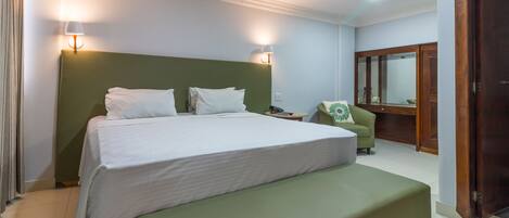 A hotel room with a large bed, a green bench, a small table, a chair, and a mirror.