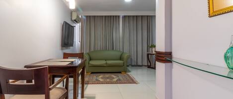 Un salón con un sofá verde, una mesa de comedor de madera y sillas. Hay un televisor montado en la pared y una estantería de cristal con un jarrón verde.