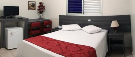 A hotel room with a bed, a desk, a chair, a small refrigerator, a bedside table with a lamp, and a window with blinds.