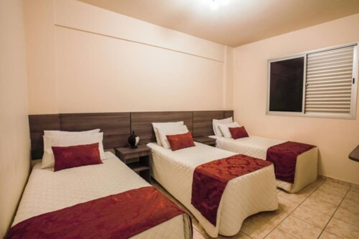 A hotel room with three single beds, each with a red blanket and white pillows. There is a window with blinds, a bedside table with a lamp, and a wall-mounted shelf.