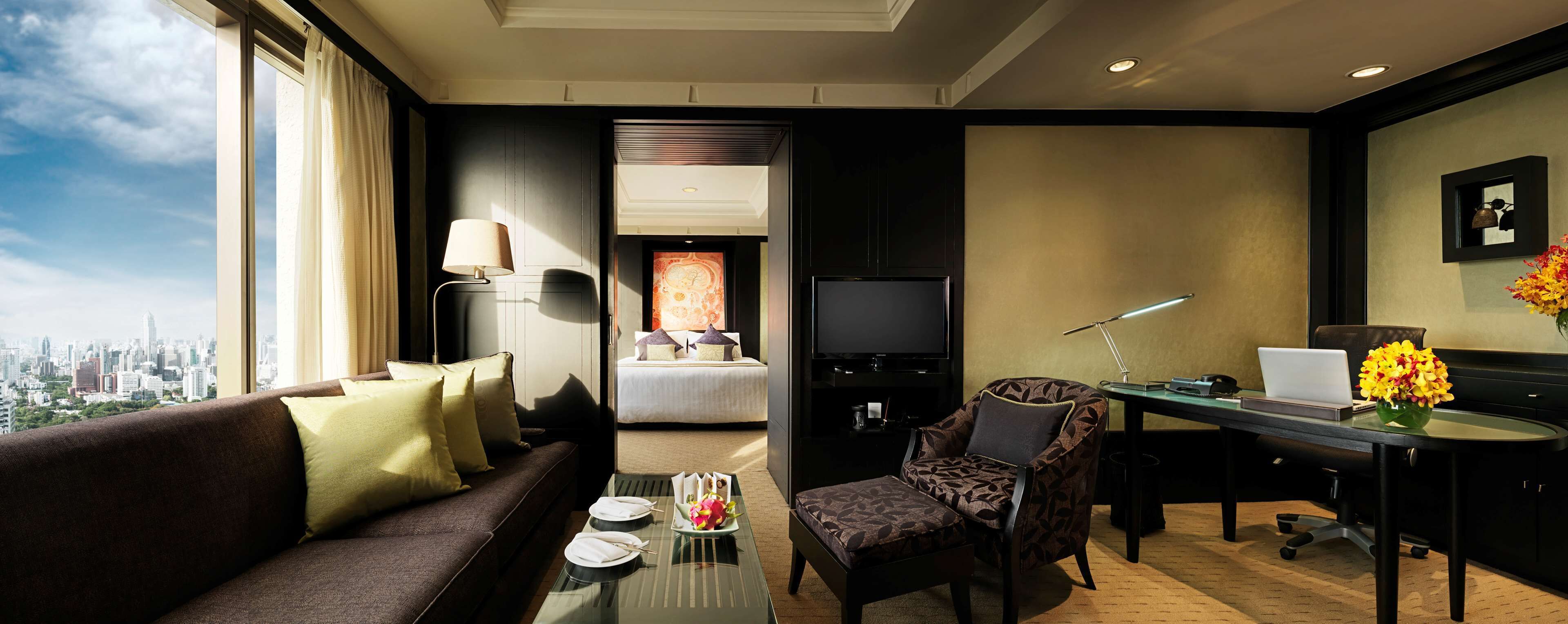 A hotel room with a sofa, a desk with a laptop, a chair, a TV, and a bed with a city view.