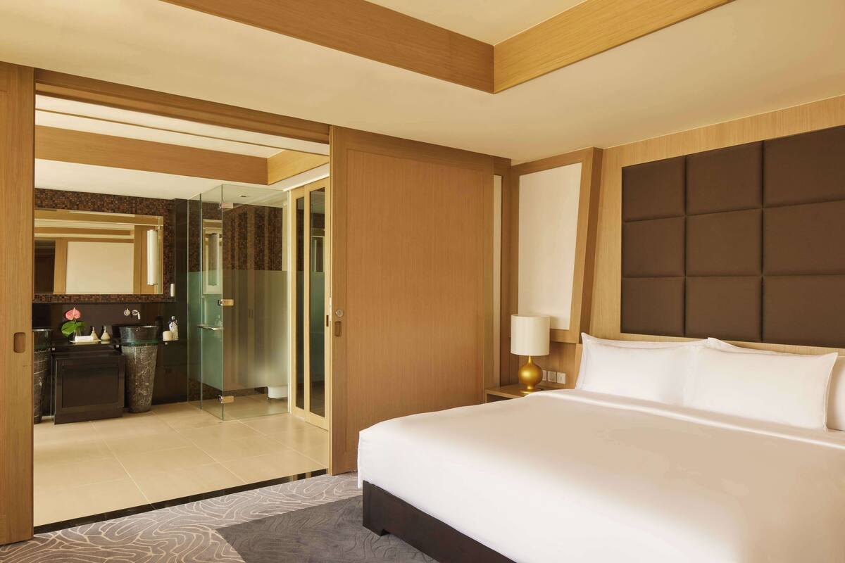 A hotel room with a large bed, a bedside table with a lamp, a glass-enclosed shower area, and a wooden door leading to a bathroom.