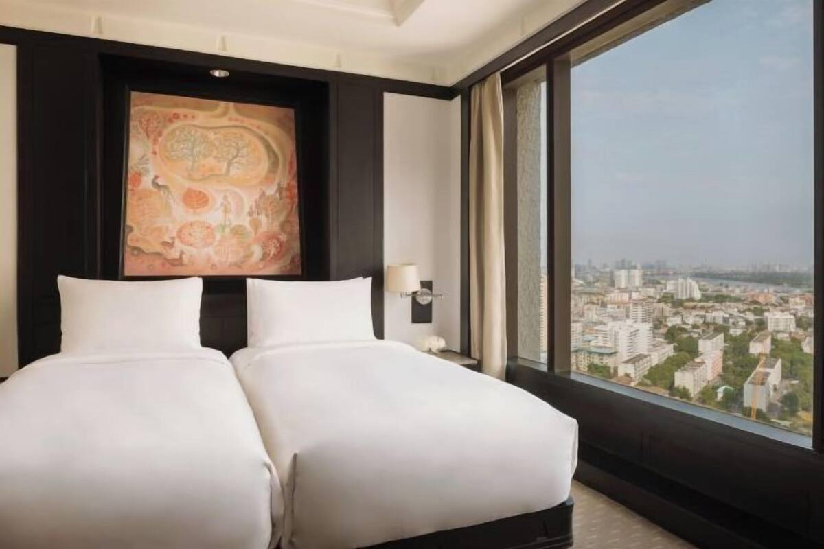 A hotel room with two beds, a large window with a city view, and a painting on the wall.