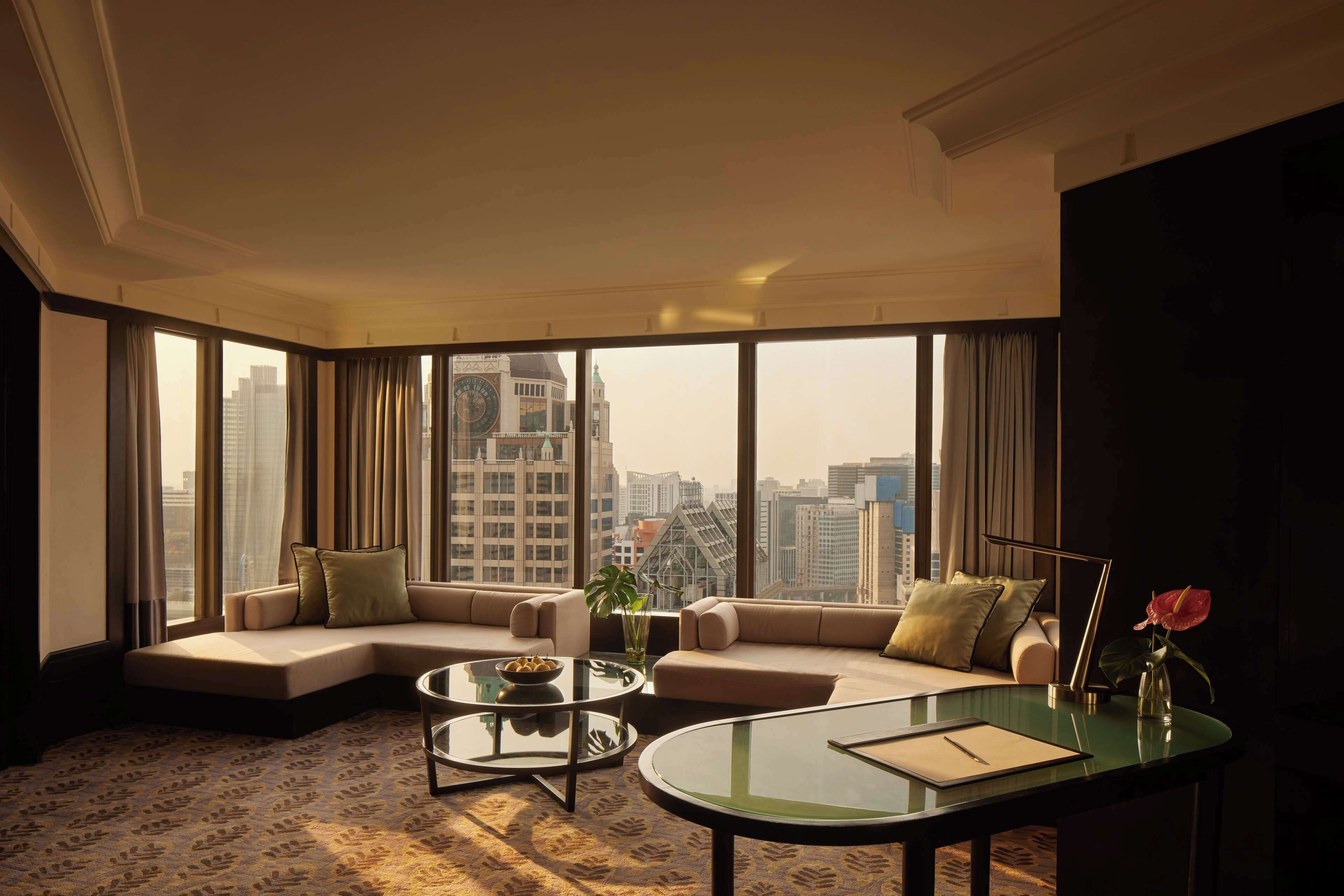 Una habitación de hotel moderna con un ventanal que ofrece vistas a la ciudad, una zona de estar acogedora con sofá y sillones, una pequeña mesa de centro y un escritorio con un jarrón de flores.