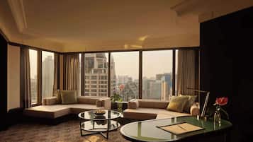 Una habitación de hotel moderna con un ventanal que ofrece vistas a la ciudad, una zona de estar acogedora con sofá y sillones, una pequeña mesa de centro y un escritorio con un jarrón de flores.