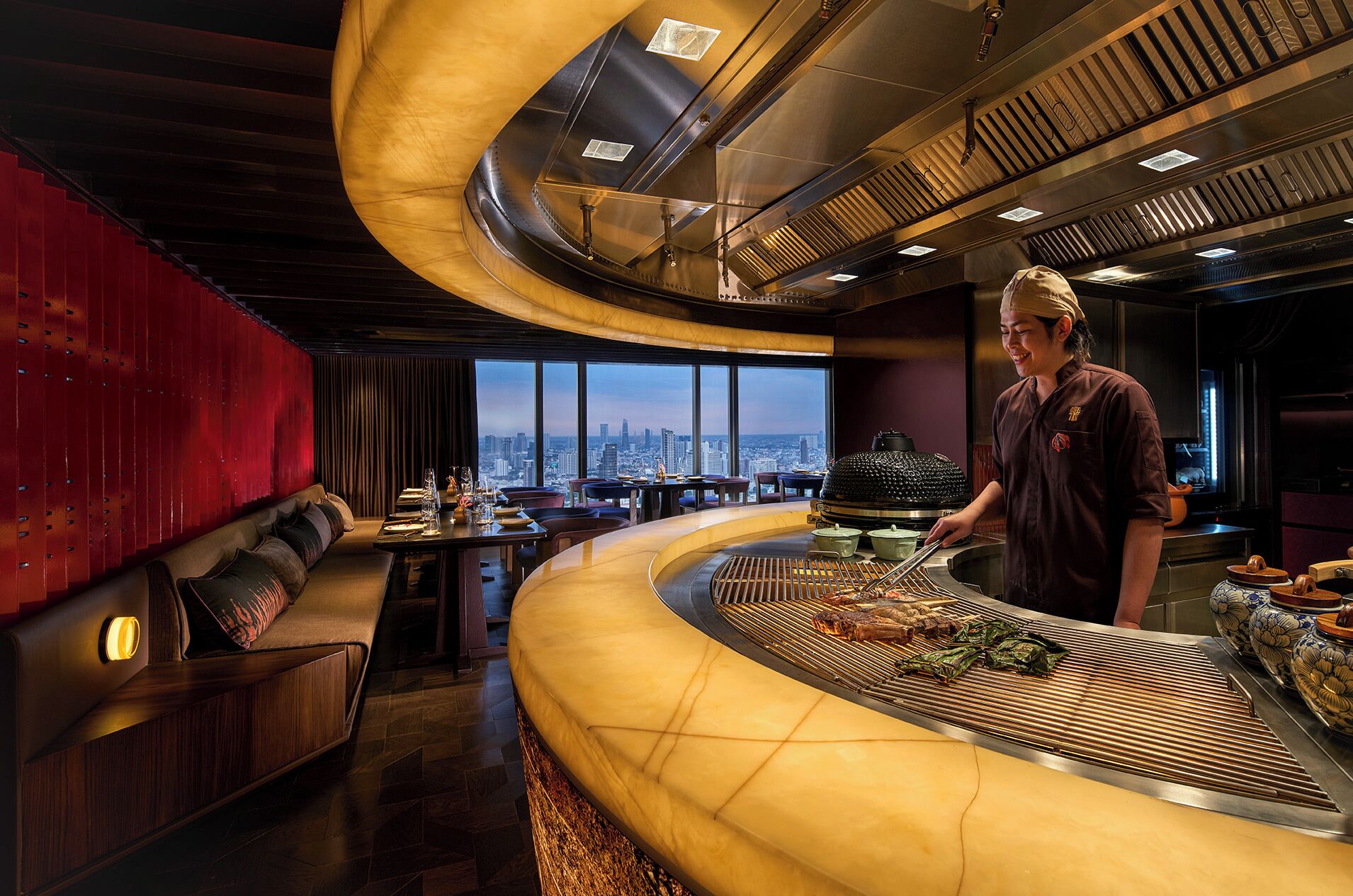 A restaurant with a curved counter, a chef preparing food, and a city view through large windows.