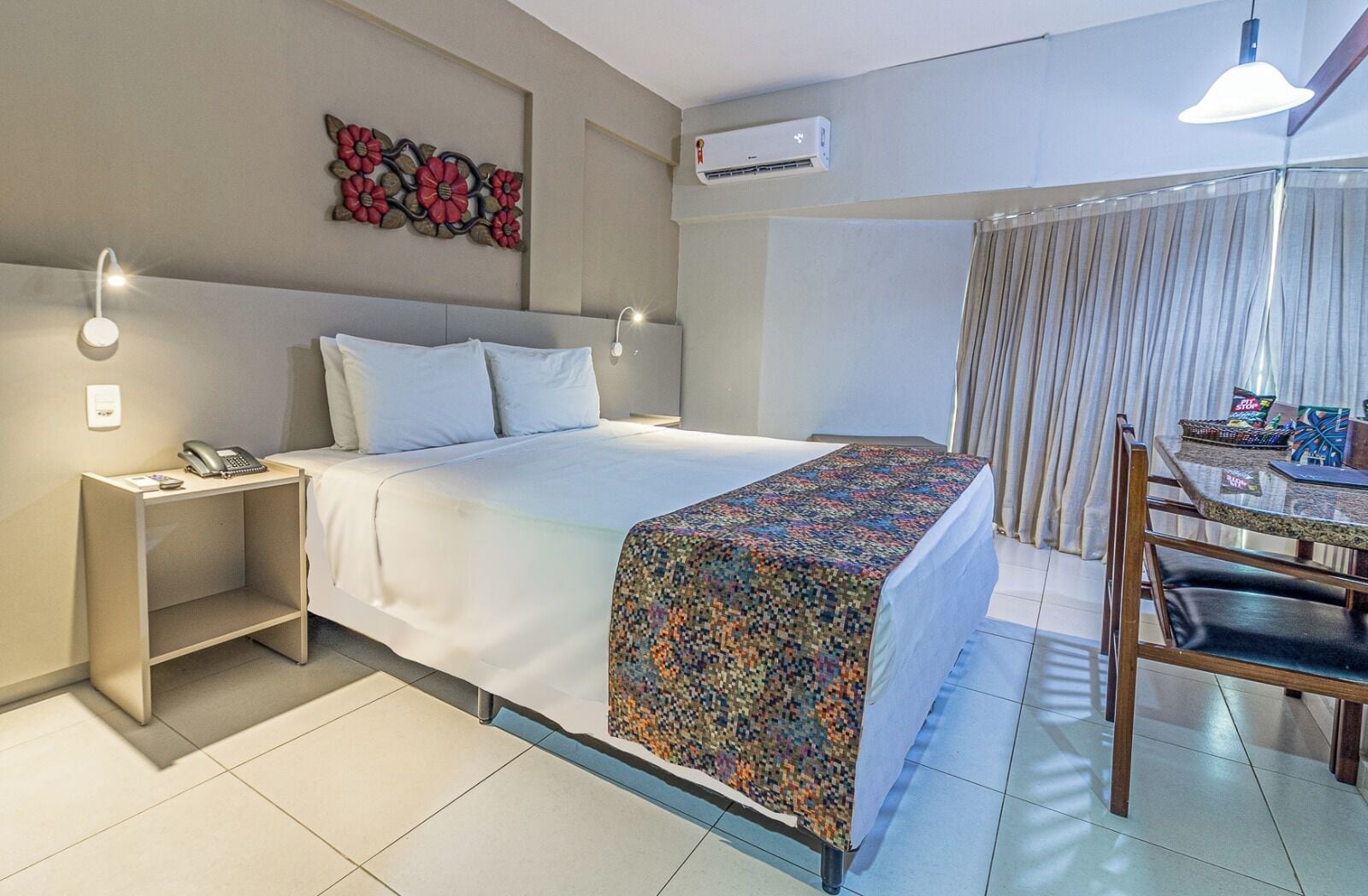 Habitació d'hotel amb un llit, una taula de nit amb un telèfon, un escriptori amb una cadira, un element decoratiu a la paret i un aparell d'aire condicionat.