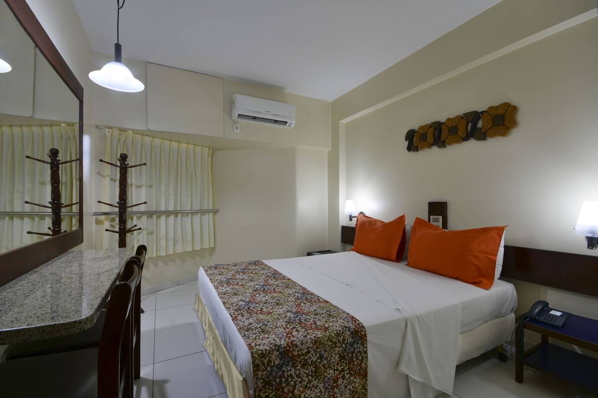 A hotel room with a bed, a desk, a chair, a mirror, and a wall-mounted air conditioner.