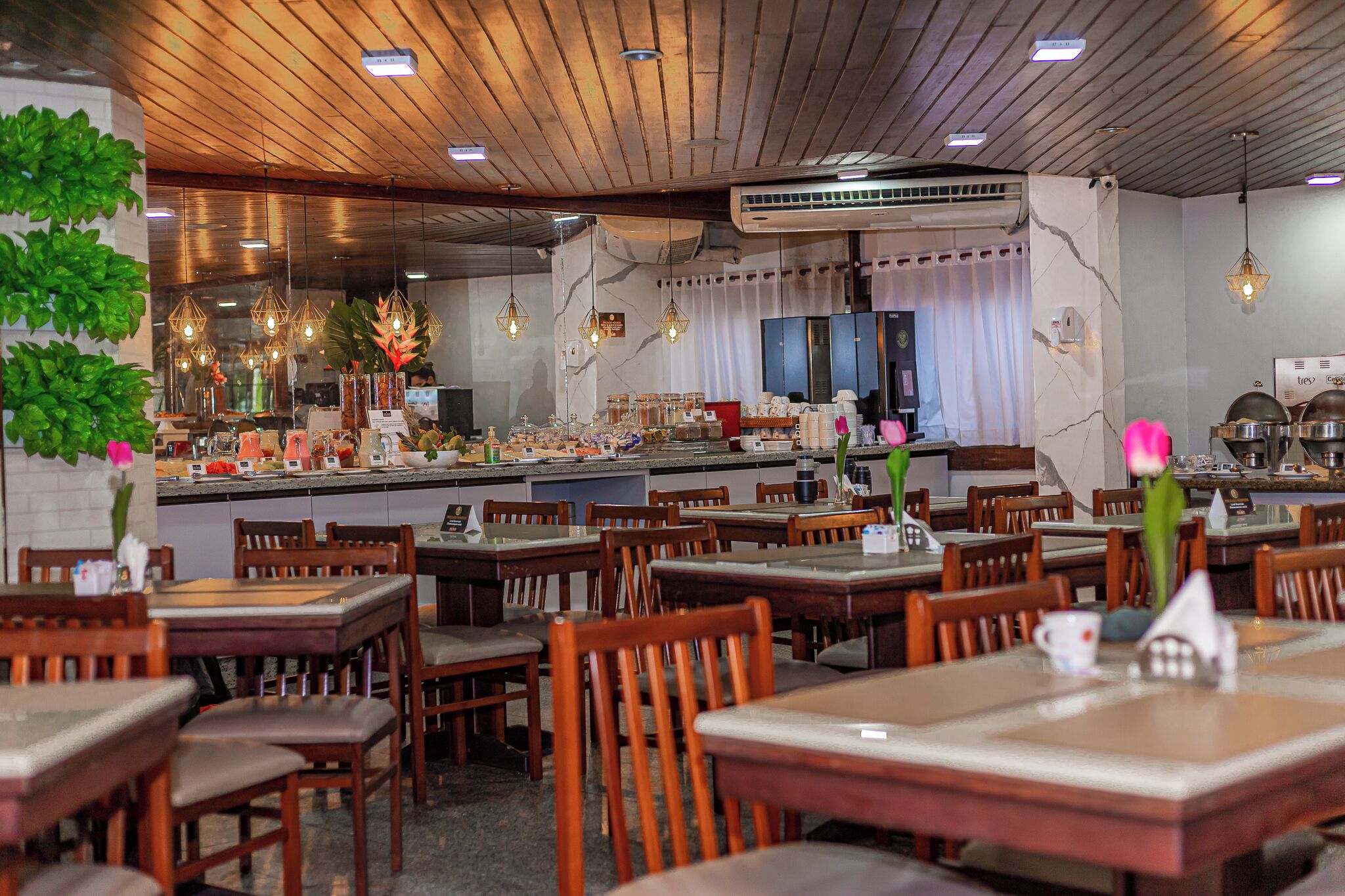 A restaurant with wooden ceiling, multiple tables and chairs, hanging lights, and a kitchen area in the background.