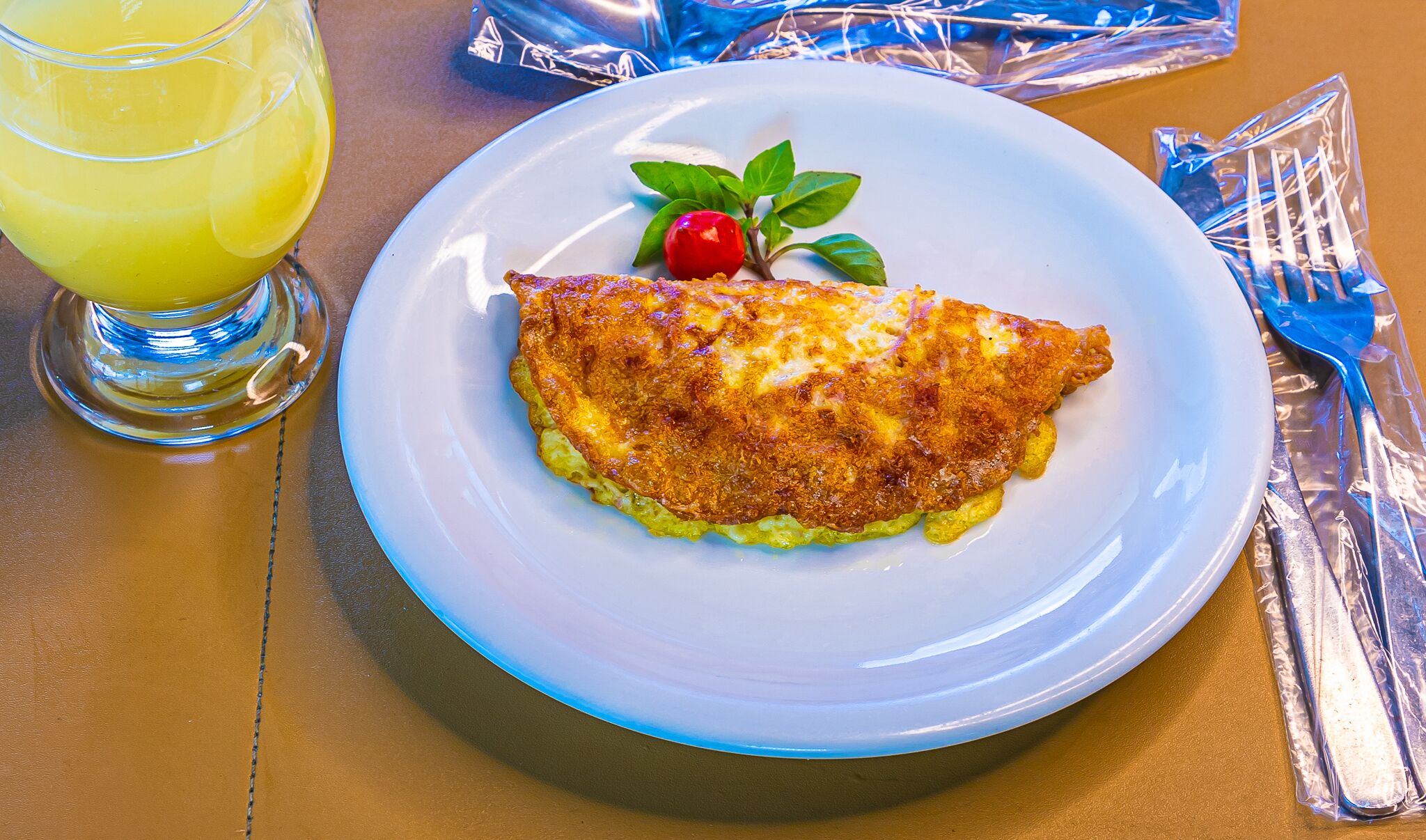 Um prato com um omelete dobrado, um copo de suco de laranja e uma cereja sobre uma folha.