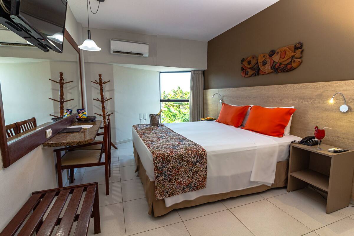 A hotel room with a bed, a nightstand, a desk with chairs, and a window with a view of greenery.