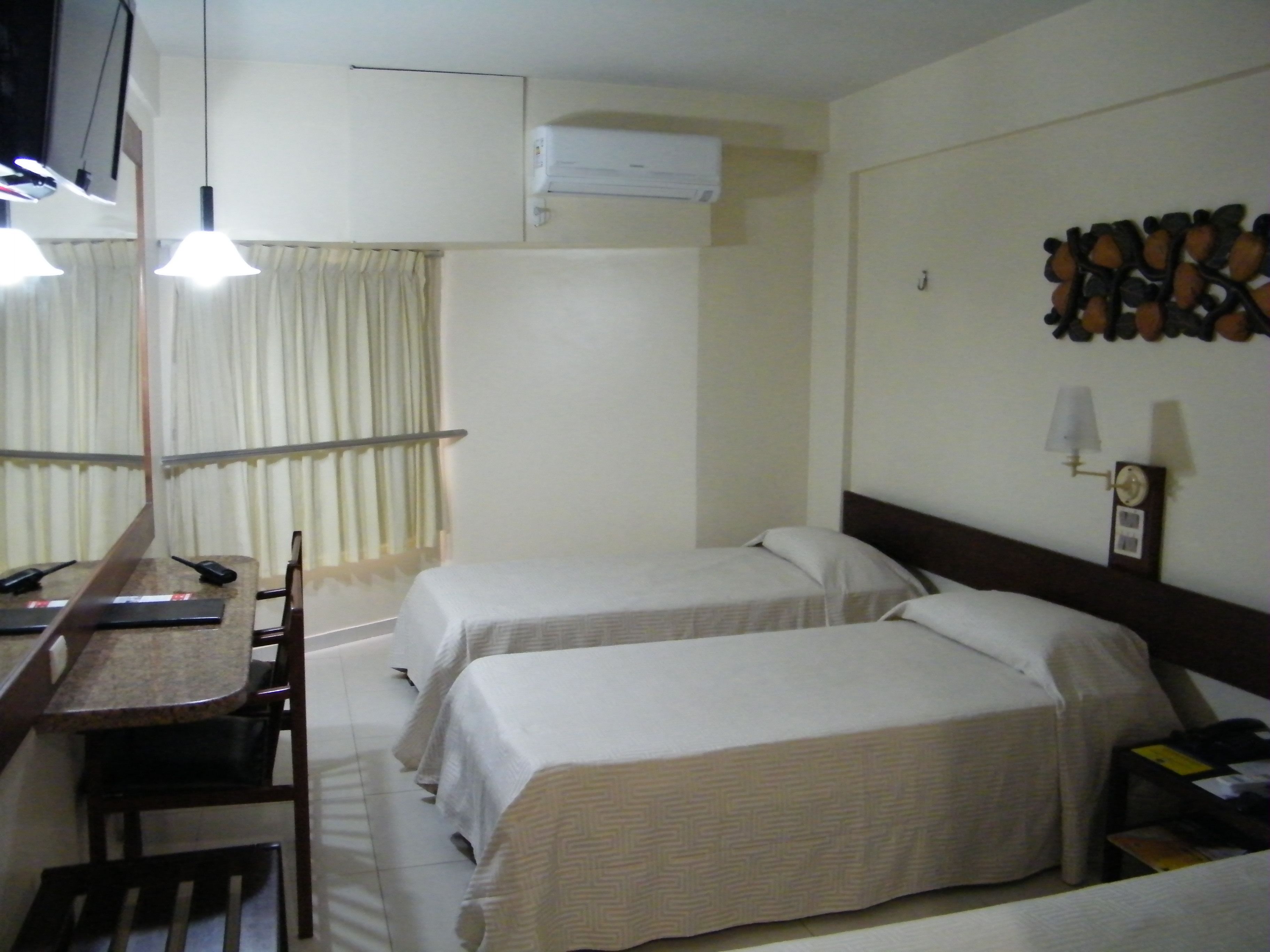 Une chambre d’hôtel avec deux lits, un bureau avec une chaise, un miroir, un climatiseur et une horloge fixée au mur.