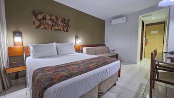 Quarto de hotel com uma cama grande, uma mesinha de cabeceira com abajur, uma pequena mesa com cadeiras e um ar-condicionado fixado na parede.