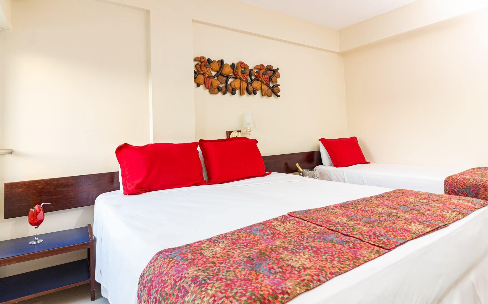 Habitación de hotel con dos camas, cada una con almohadas rojas y una colcha blanca. Hay una manta tejida de colores en el suelo. Se observa una decoración mural con pájaros sobre las camas.