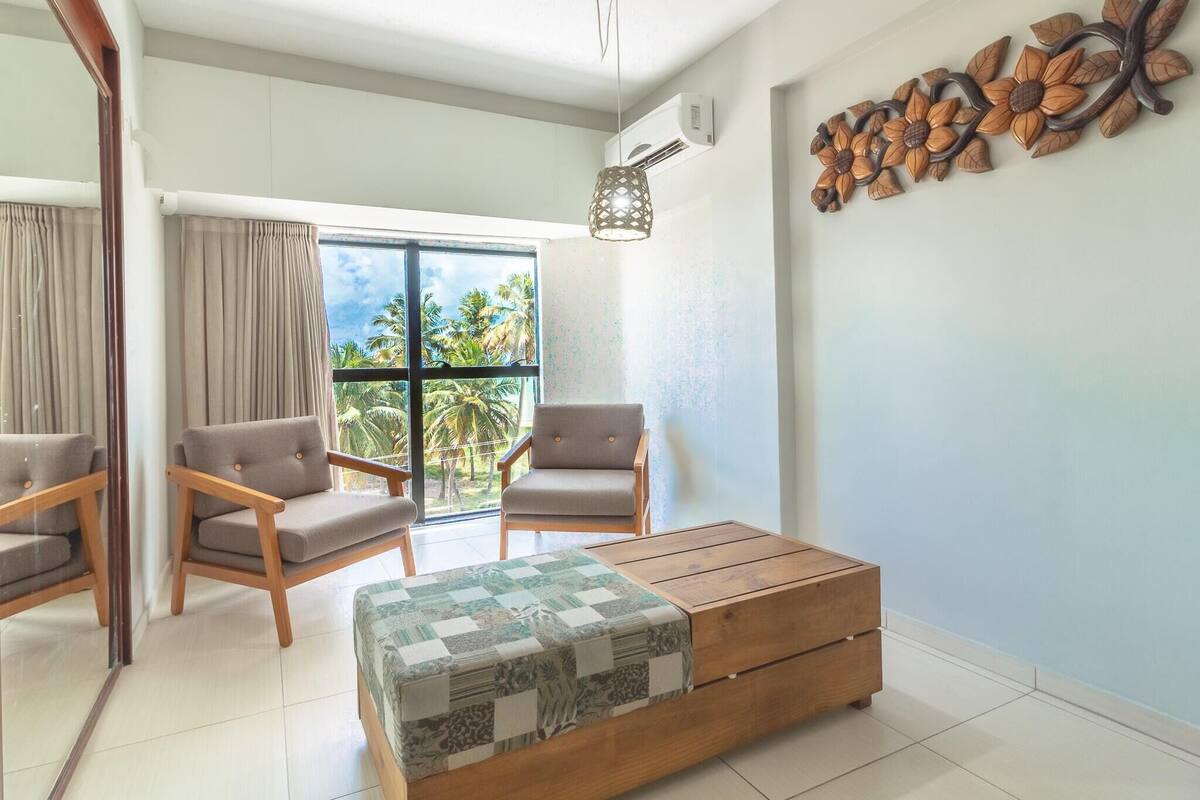 A modern living room with a wooden sofa, two armchairs, a large window with a view of palm trees, and a decorative wall piece.