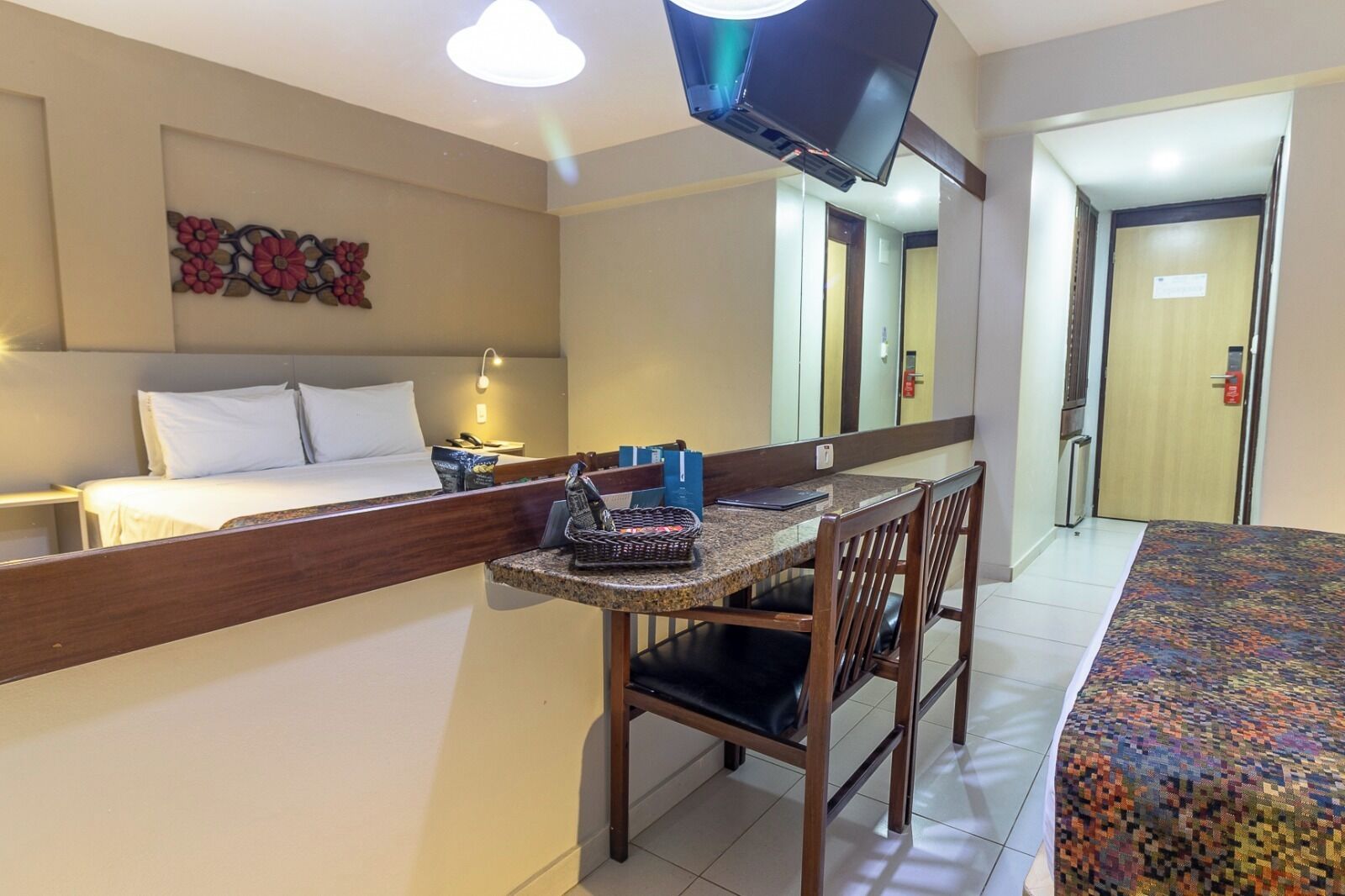 Una habitación de hotel con una cama, un escritorio con silla, un televisor montado en la pared y una puerta al fondo.