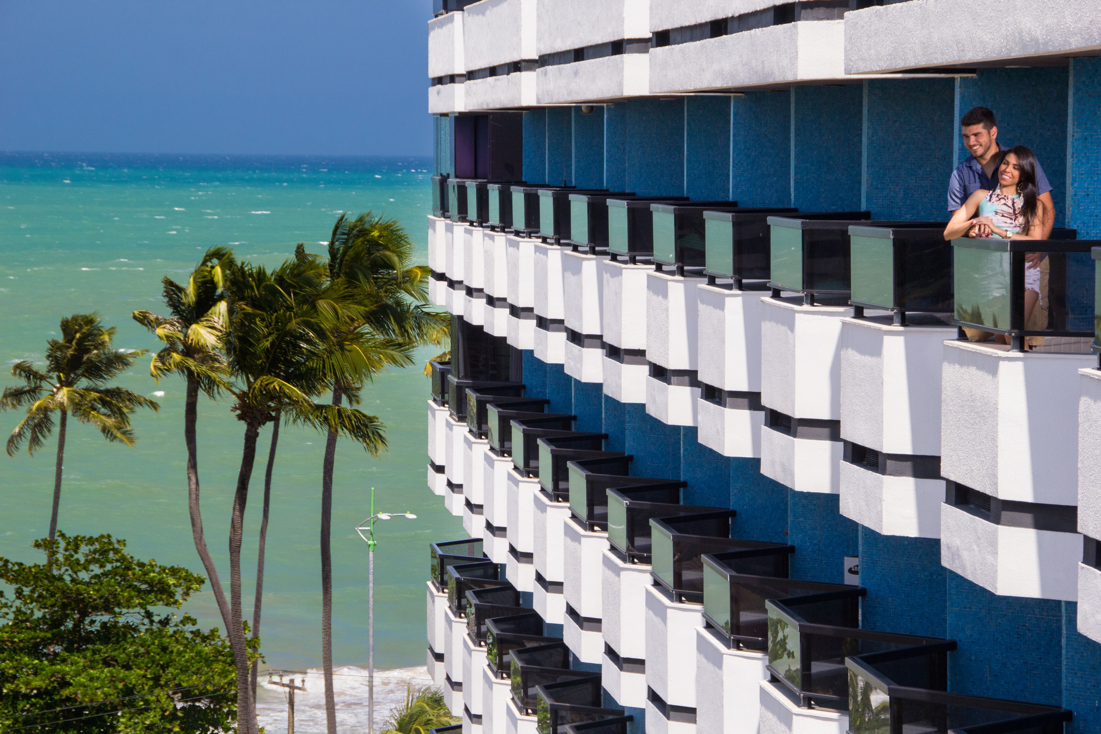 Et hotel med moderne design, med altaner der har udsigt til havet, palmer og en klar blå himmel.