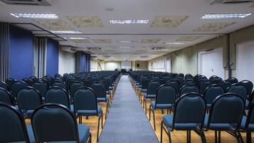 Un salón de conferencias con filas de sillas, piso alfombrado y una pantalla de proyector al frente.