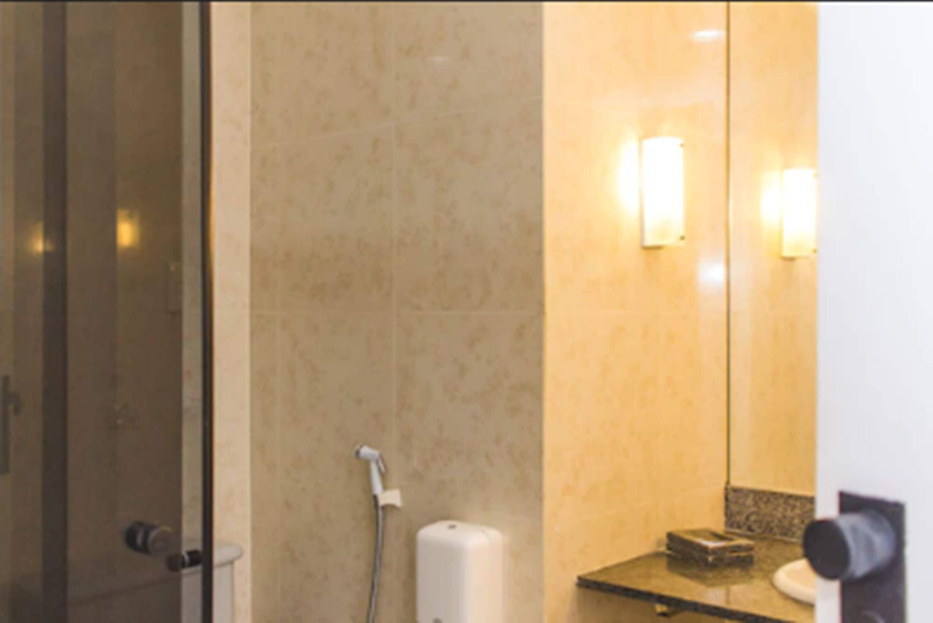 Un baño con mampara de vidrio, secador de manos y espejo con luces empotradas.