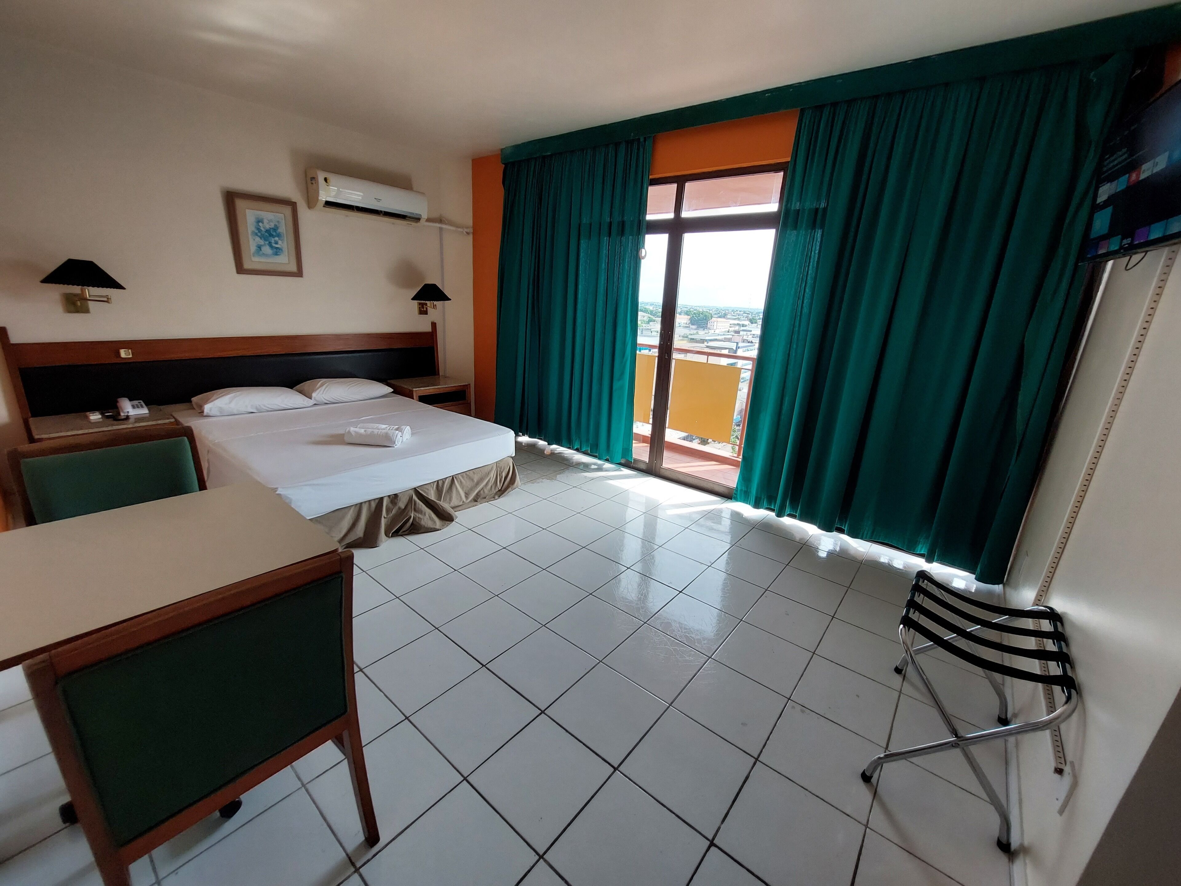 A hotel room with a bed, a desk, a chair, a TV, and a balcony with curtains.