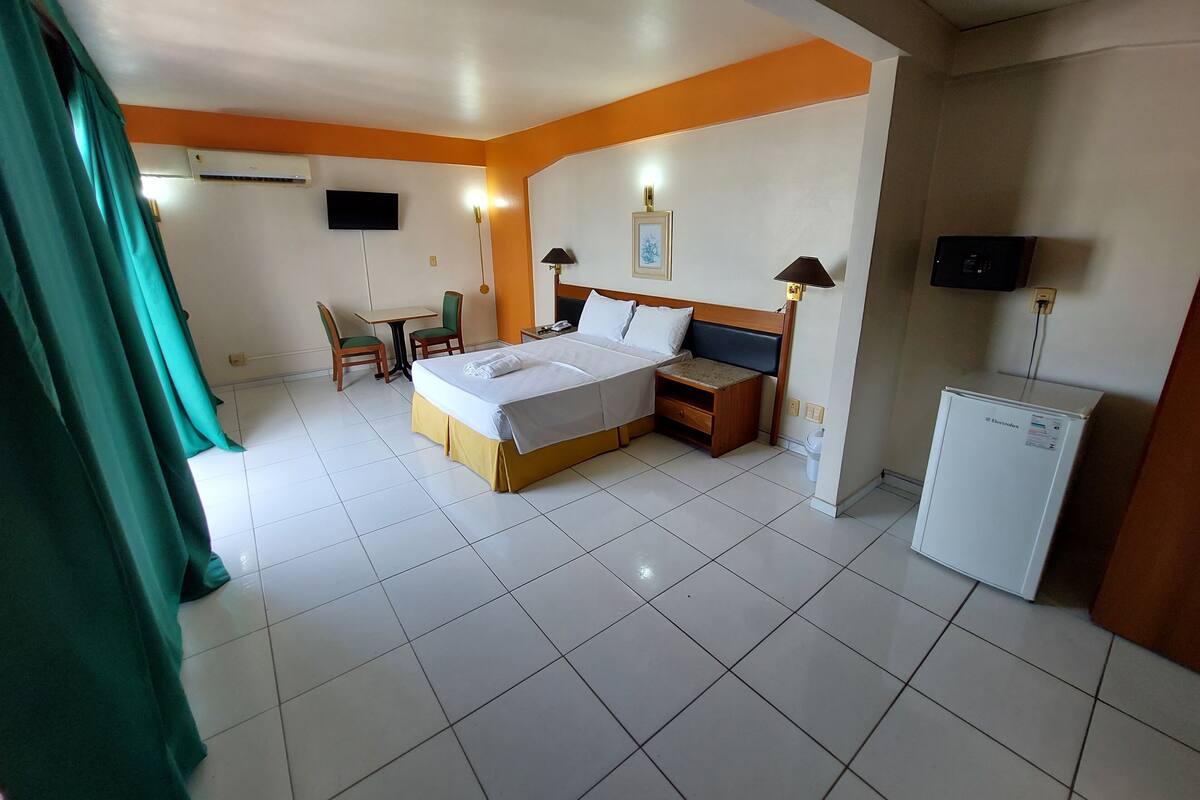 A hotel room with a bed, a desk, a chair, a TV, a lamp, and a refrigerator.