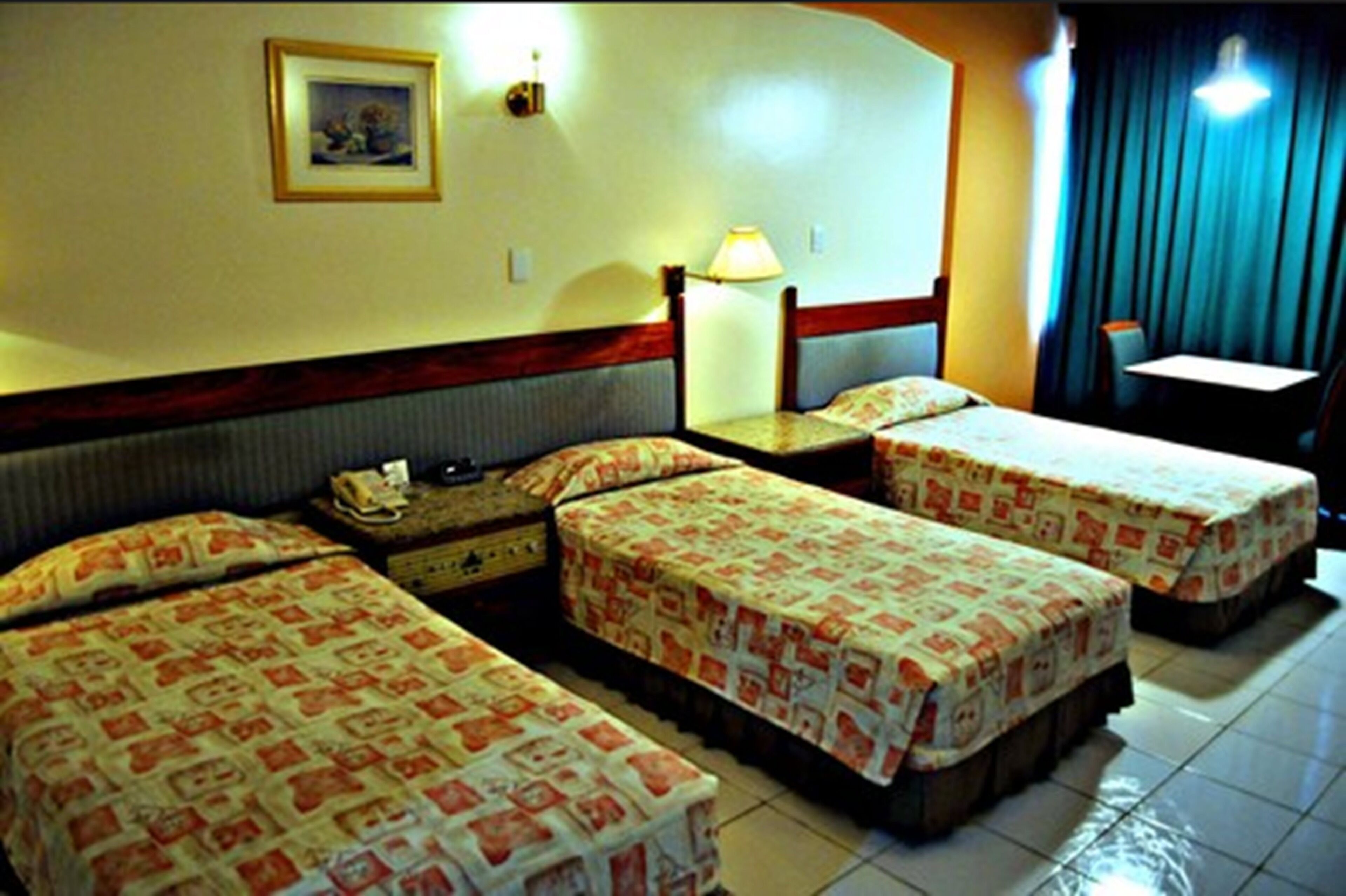 A hotel room with three single beds, a small desk, a chair, and a lamp.