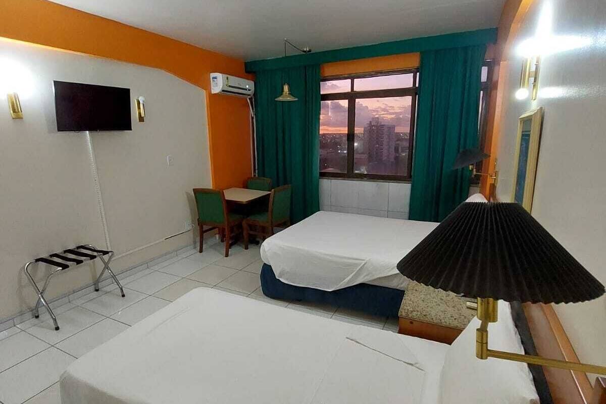 A hotel room with two beds, a TV, an air conditioning unit, a small table with chairs, and a window with curtains.