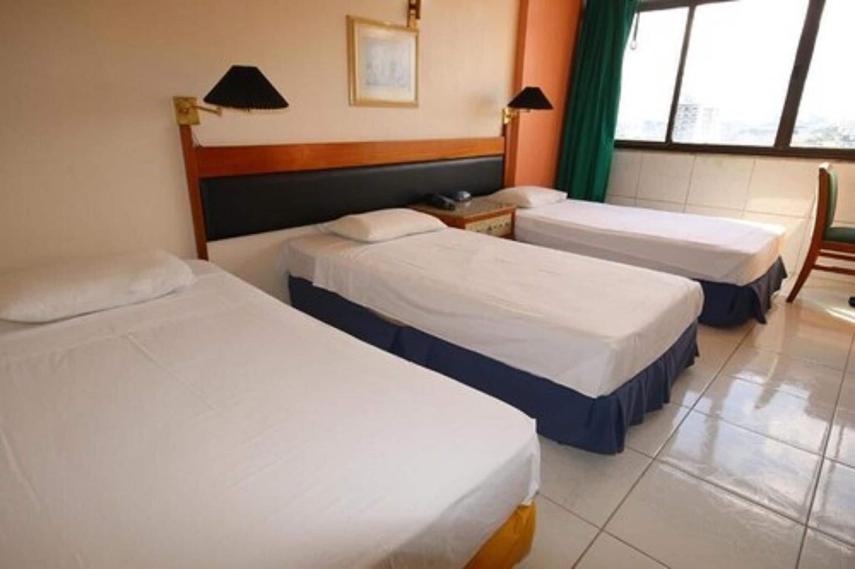 Une chambre d’hôtel avec trois lits simples, une tête de lit en bois, deux lampes de chevet, une petite table et une fenêtre avec des rideaux.