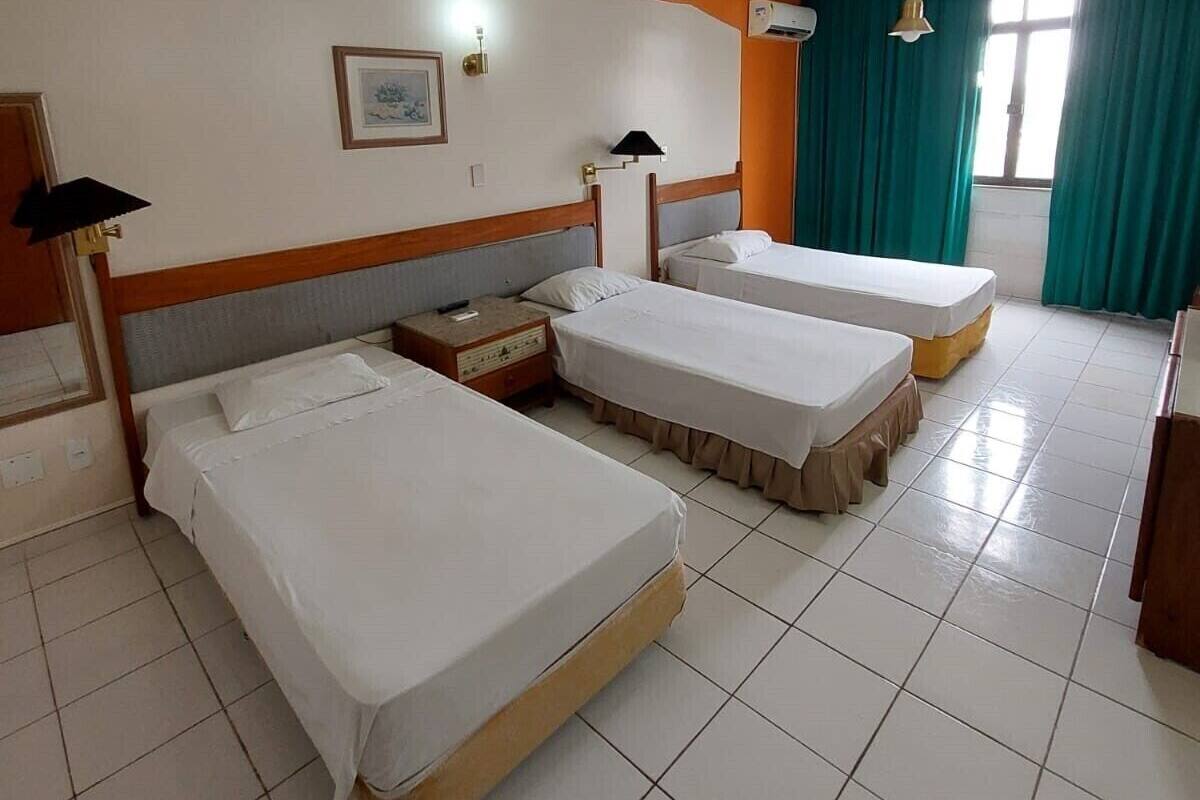 A hotel room with three single beds, a wooden headboard, a nightstand, a framed picture, and teal curtains.