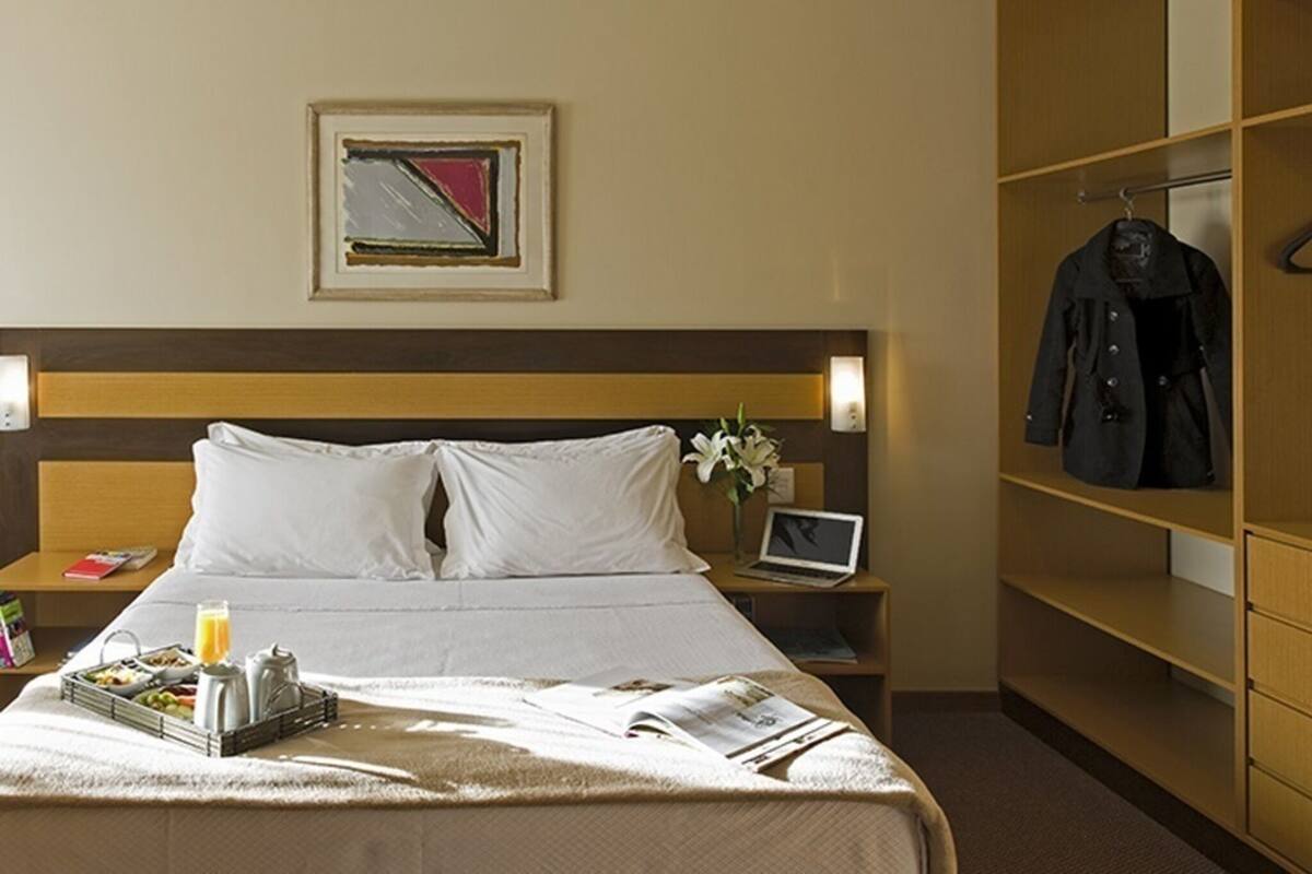 Ein Hotelzimmer mit Bett, Nachttischen, einem Laptop, einem Kleiderständer und einem gerahmten Bild an der Wand.