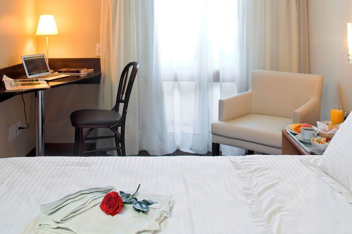 Quarto de hotel com cama, escrivaninha com laptop, cadeira, mesa de jantar com café da manhã e janela com cortinas.