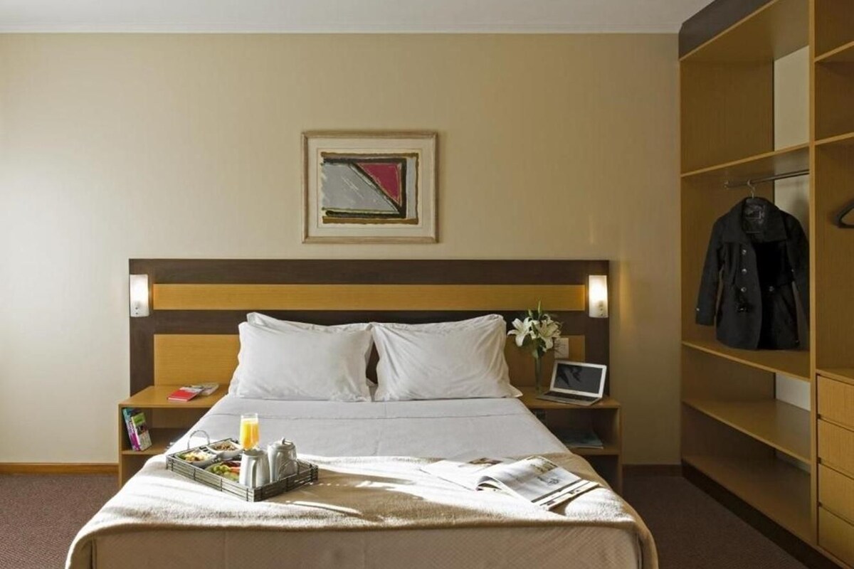 Quarto de hotel com cama, mesas de cabeceira, um laptop, um quadro emoldurado e um porta-casacos.