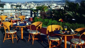 A terrace with wicker chairs and tables, overlooking a cityscape with buildings and hills. - Piazza Navona Porto Alegre by Intercity (Porto Alegre)