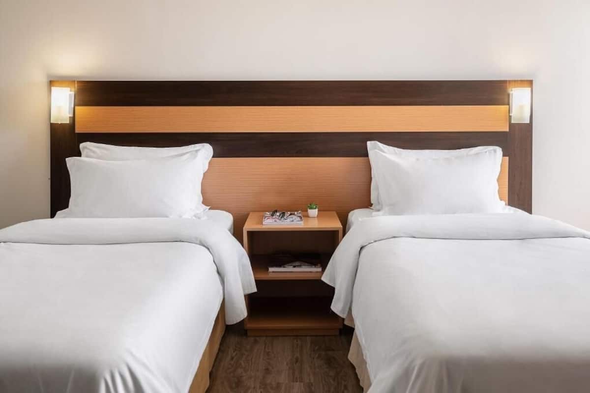 Ein Hotelzimmer mit zwei Betten, einem Holzkopfteil, Nachttischen mit Lampen und einem kleinen Regal mit einer Pflanze.