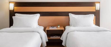 Habitación de hotel con dos camas, cabeceros de madera, mesitas de noche con lámparas y un pequeño estante con una planta.