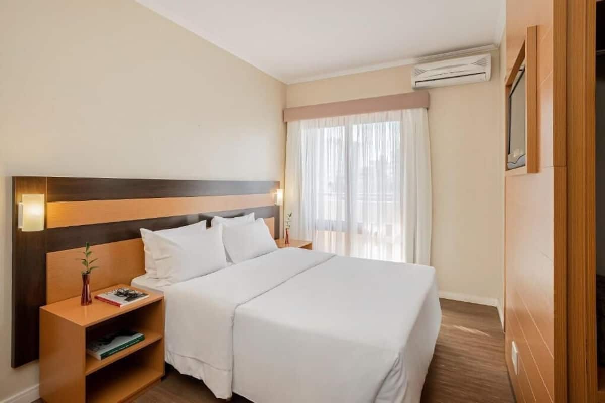 Ein Hotelzimmer mit einem großen Bett, einem Holzkopfteil, einem Nachttisch mit Lampe und einem Fenster mit durchscheinenden Vorhängen.