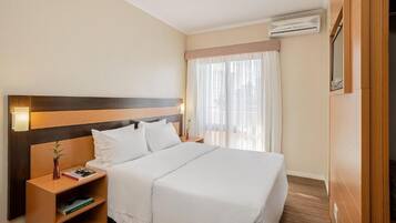Une chambre d’hôtel avec un grand lit, une tête de lit en bois, une table de chevet avec une lampe et une fenêtre ornée de rideaux légers.