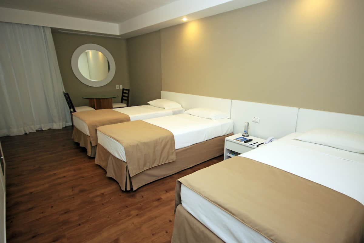 Habitación de hotel con tres camas, suelo de madera, un espejo redondo y una pequeña mesa con sillas.