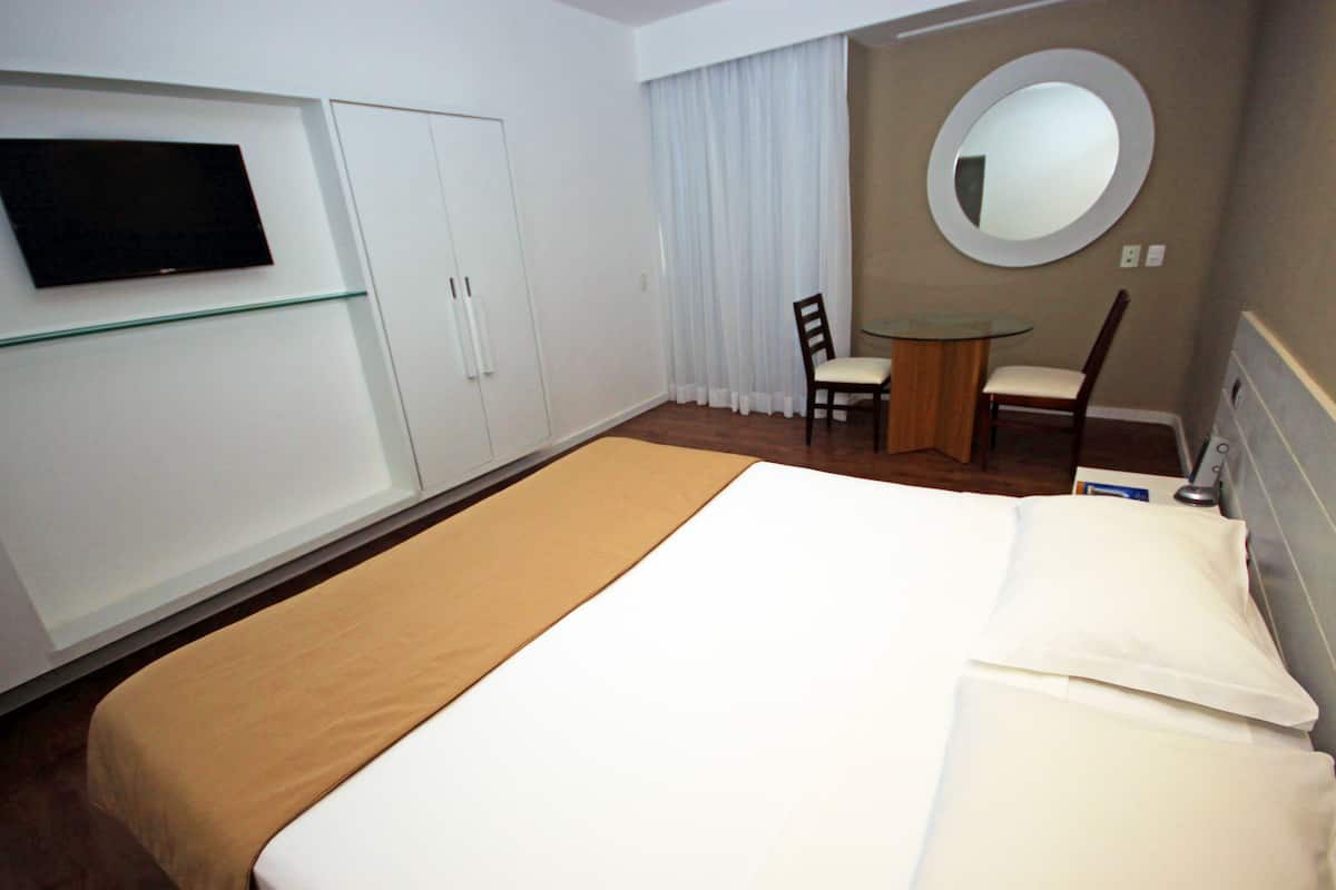 Habitación de hotel con una cama, un televisor montado en la pared, una mesita con una silla y un espejo redondo en la pared.