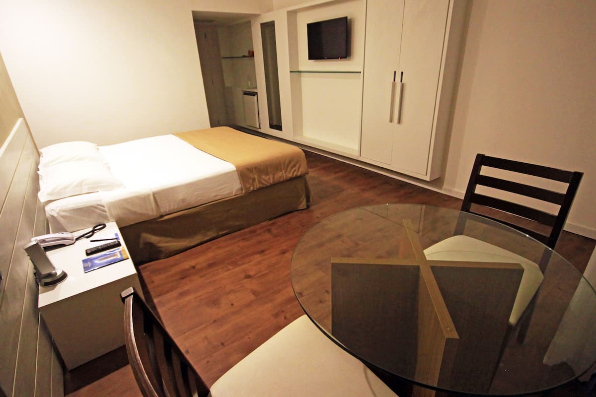 Una habitación pequeña y funcional con cama, escritorio con silla, una mesita de vidrio y televisor montado en la pared.
