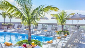 Zona de piscina con sillones de descanso, sombrillas y una mesa con alimentos para el desayuno.