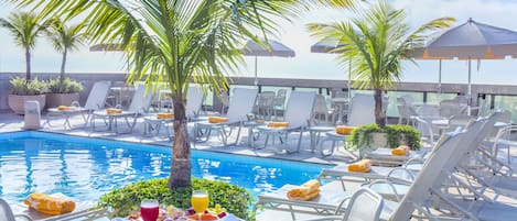 Un espace aménagé au bord de la piscine, avec des chaises longues, des parasols et une table où sont servis les articles du petit-déjeuner.