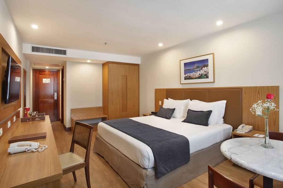 A hotel room with a bed, a desk, a chair, a small table, and a TV.