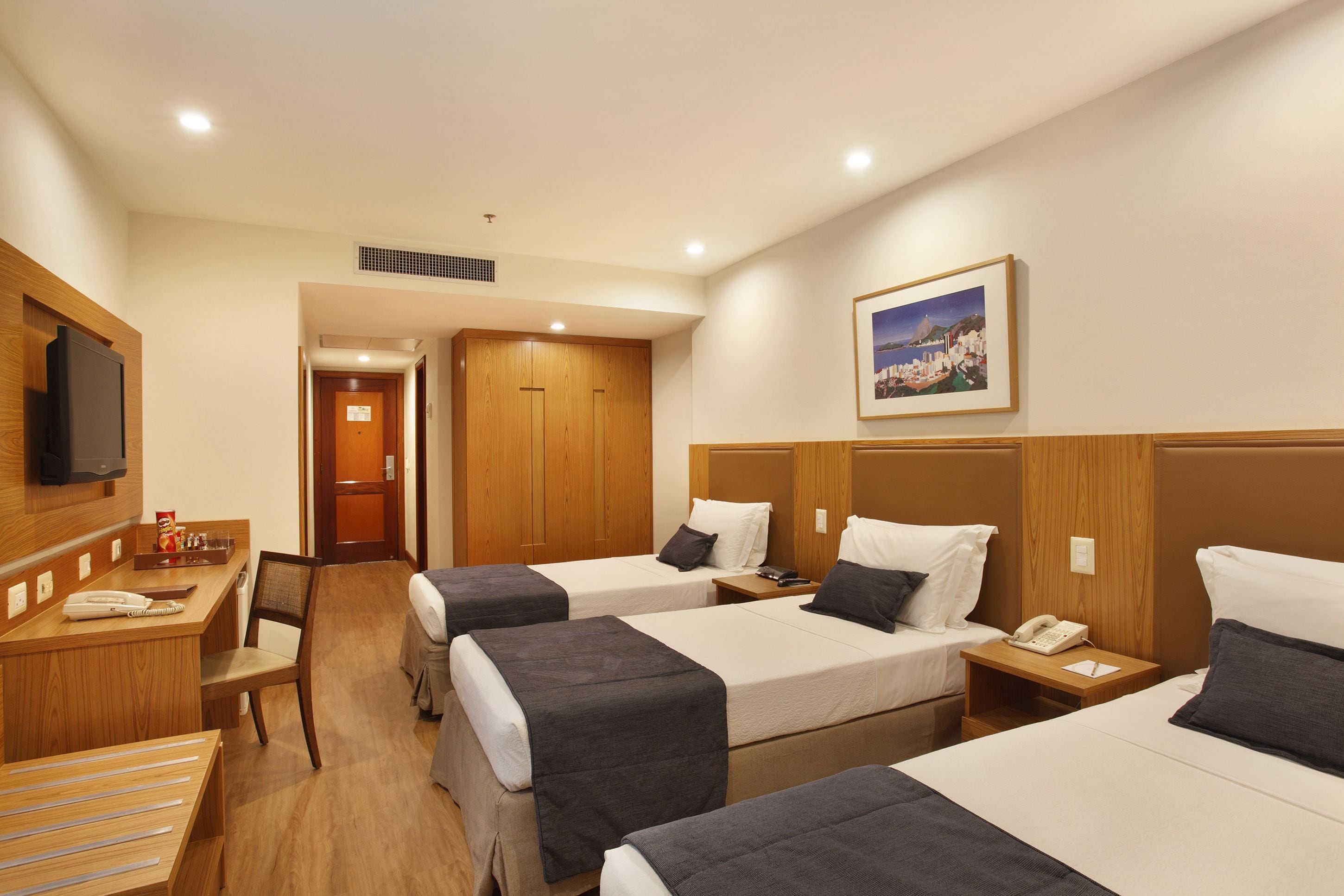 A hotel room with three beds, a desk, a chair, a TV, and a painting on the wall.