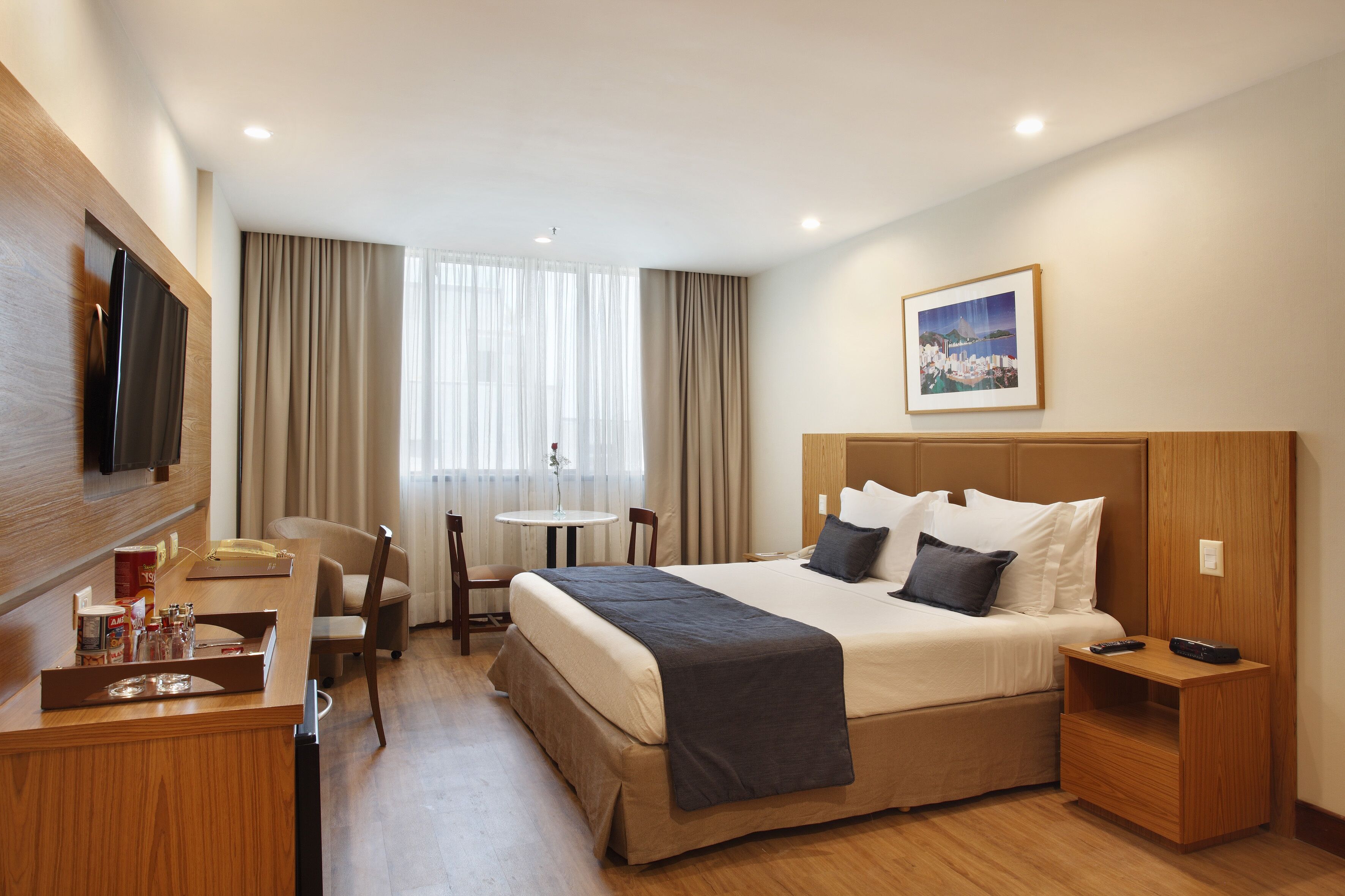 Quarto de hotel com cama, mesa de cabeceira, televisão, escrivaninha com cadeira e uma pequena mesa com uma cadeira.